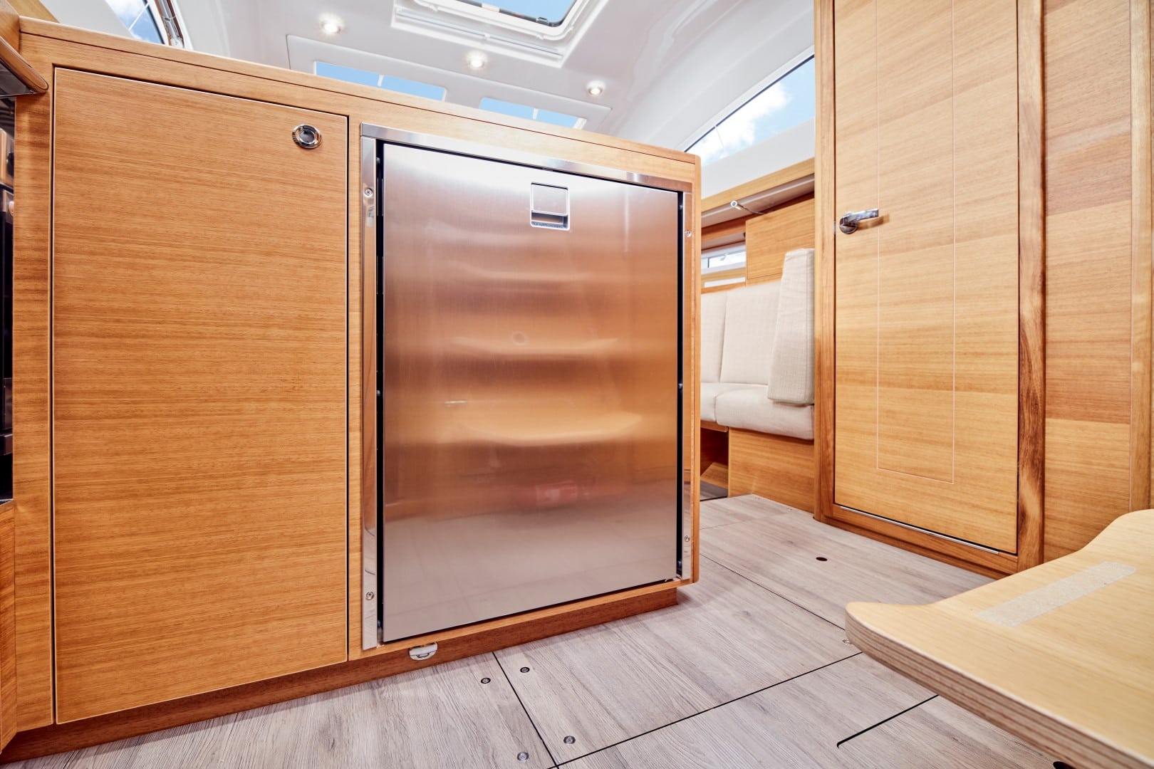Innenansicht eines modernen Wohnmobils mit einer Glasschiebetür zwischen Holzschränken, die zu einer gemütlichen Sitzecke mit beiger Polsterung auf einer Segelyacht führt.