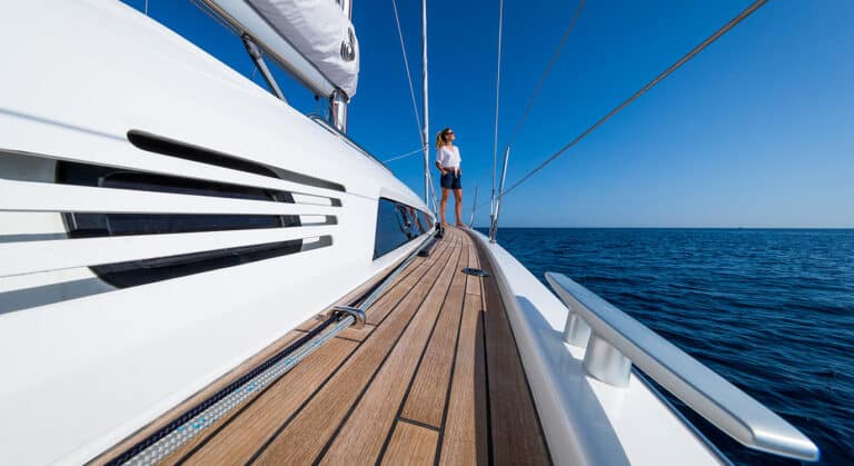 Eine Person steht am Bug einer eleganten weißen Segelyacht und blickt auf den Ozean, mit klarem blauen Himmel darüber und ruhigem blauen Wasser rundherum.