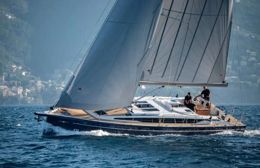 Ein schnittiger Katamaran mit großen Segeln gleitet über einen ruhigen, klaren See, umgeben von einer Berglandschaft unter einem sonnigen Himmel, und an Deck sind ein paar Menschen zu sehen.