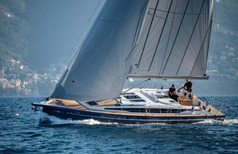 Ein schnittiger Katamaran mit großen Segeln gleitet über einen ruhigen, klaren See, umgeben von einer Berglandschaft unter einem sonnigen Himmel, und an Deck sind ein paar Menschen zu sehen.