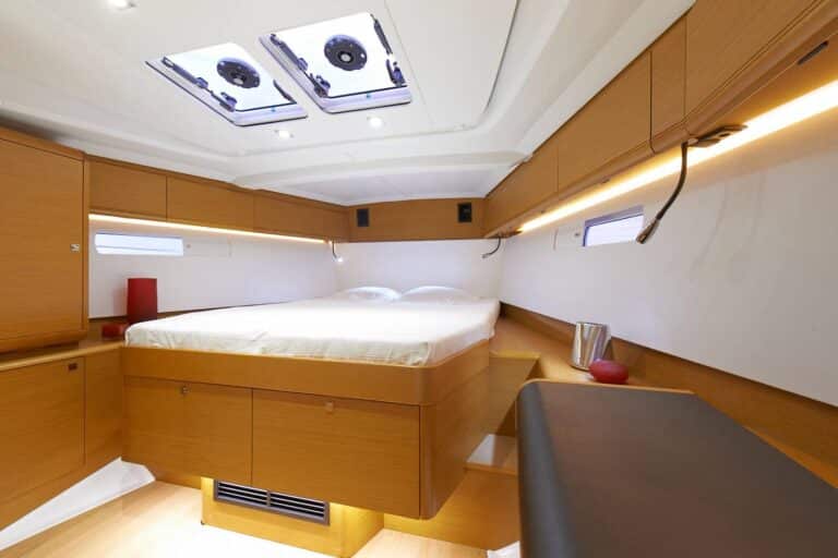 Innenansicht einer Luxus-Segelyachtkabine mit großem Bett, Holzschränken und Dachluken für Tageslicht. Helles und modernes Design mit gemütlicher Sitzecke auf der rechten Seite.