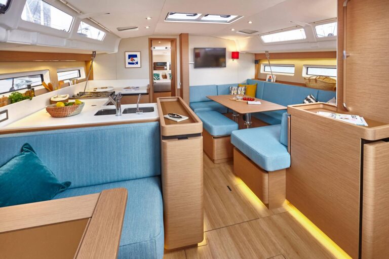 Innenansicht einer modernen Yachtkabine mit geräumigem Design mit Holzakzenten, einem blau gepolsterten U-förmigen Essbereich und natürlichem Licht, das während eines Segeltörns durch mehrere Fenster einfällt.