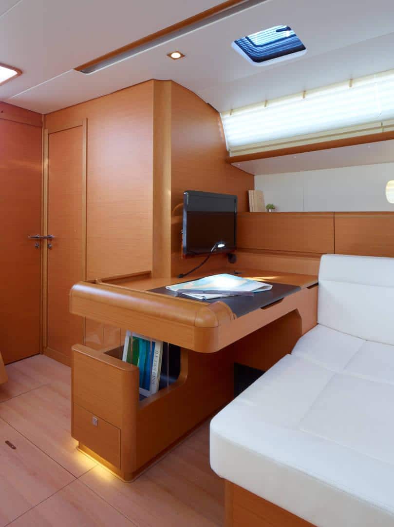 Eine moderne Yachtkabine mit einem ordentlich organisierten Arbeitsbereich mit Holzschreibtisch, Computermonitor und weißem Plüschsofa. Natürliches Licht strömt durch ein Seitenfenster ein und unterstreicht die warmen Innentöne des Katamarans.