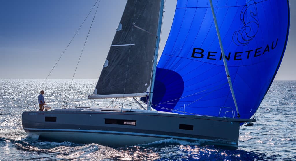 Ein Segelboot mit einem auffälligen blauen Spinnaker mit der Aufschrift „BENETEAU“ segelt auf einem glitzernden Ozean. Ein Mann steht auf dem Bug und blickt in die Ferne. Leichte Wellen auf dem Wasser reflektieren das helle Sonnenlicht während ihres Segelurlaubs.