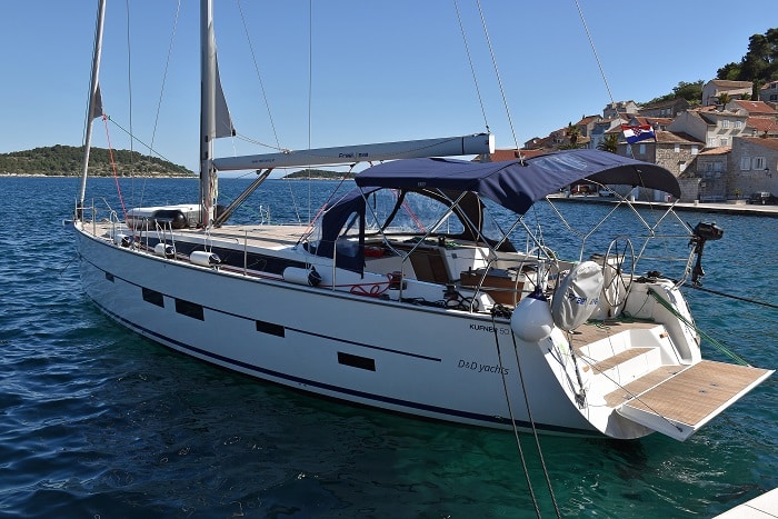 Eine weiße Segelyacht liegt unter klarem blauen Himmel in der Nähe einer Küstenstadt vor Anker. Die Yacht hat ein marineblaues Biminitop, Teakdecks und heißt „Kunigunda“. Sie ist für Mitsegeln konzipiert.