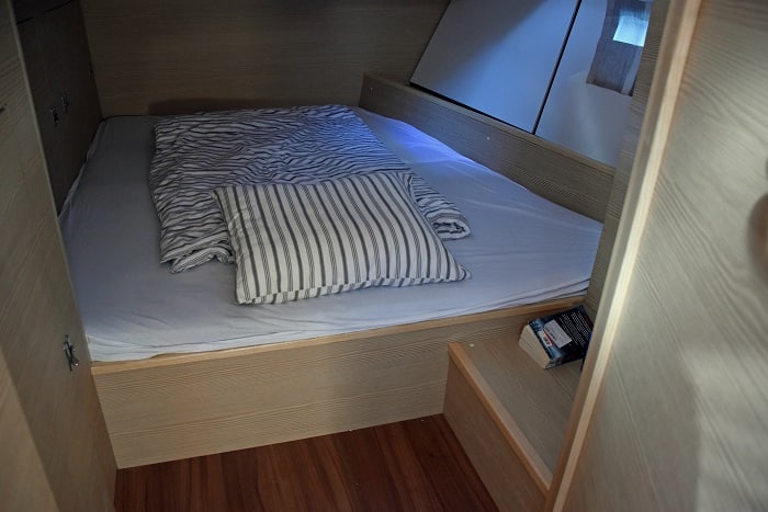 Ein ordentlich gemachtes Bett in einem kompakten, holzgetäfelten Zimmer mit gestreifter Bettwäsche und einem kleinen Nachttischregal mit Büchern, das während eines Segeltörns vom natürlichen Licht eines Fensters beleuchtet wird.