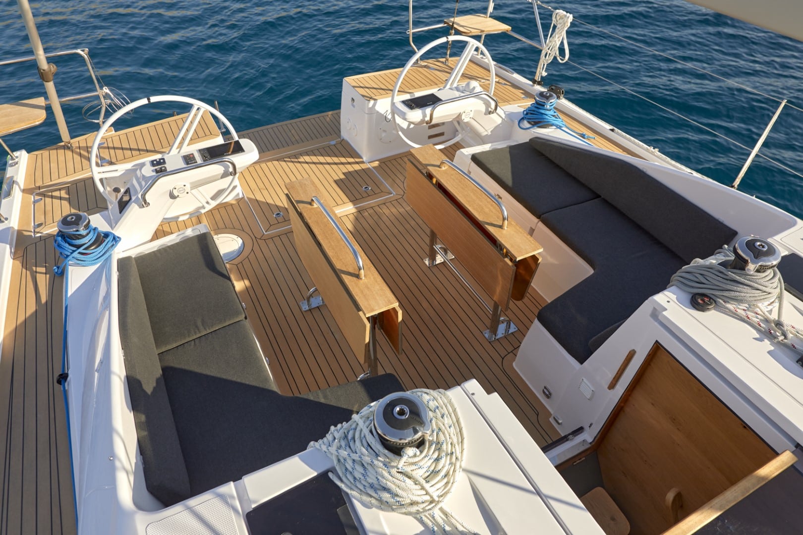 Ein Yachtdeck mit Holzboden, zwei Steuerrädern und gepolsterten Sitzbereichen unter einem klaren blauen Himmel auf einem Katamaran.