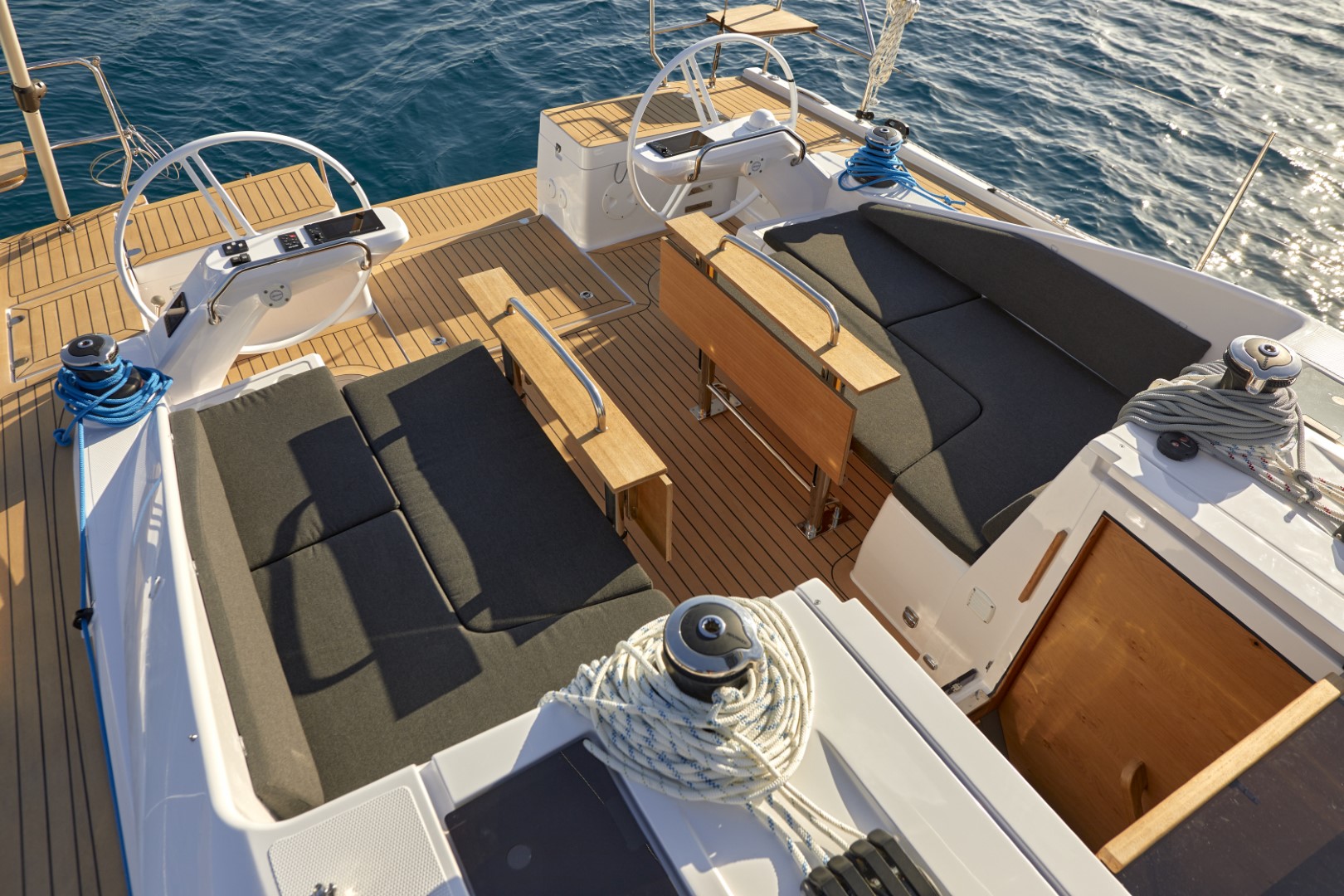Rückansicht des Decks eines Luxus-Katamarans mit Holzboden, Plüschsitzen, Steuerrad und Segelausrüstung unter klarem blauen Himmel auf ruhigem Meer.