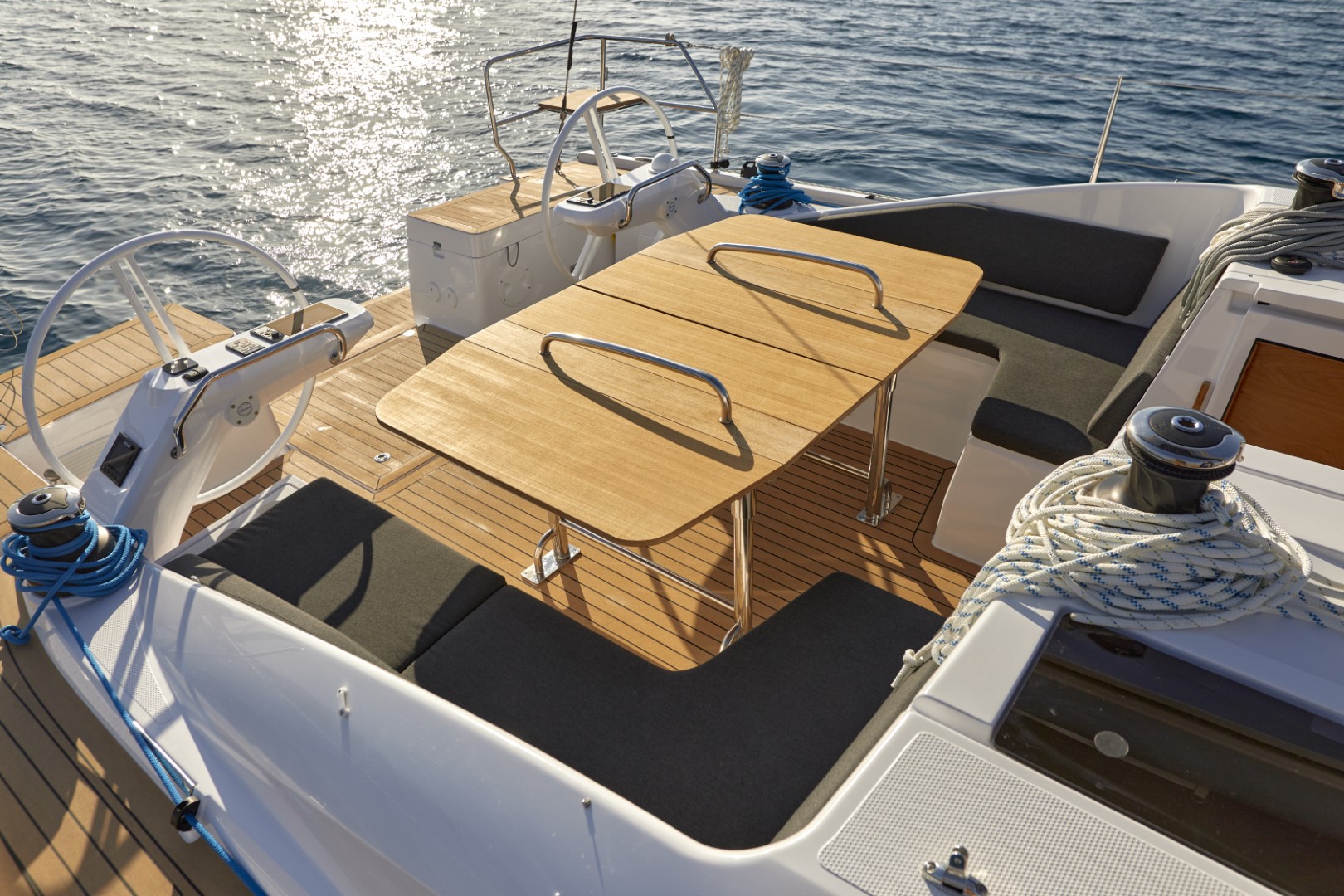 Ein luxuriöses Yachtdeck mit zwei hölzernen Steuerrädern auf beiden Seiten, ausgestattet mit einem Holztisch und gepolsterten Sitzgelegenheiten, sonnendurchflutet auf glitzerndem Wasser während eines idyllischen Segelurlaubs.