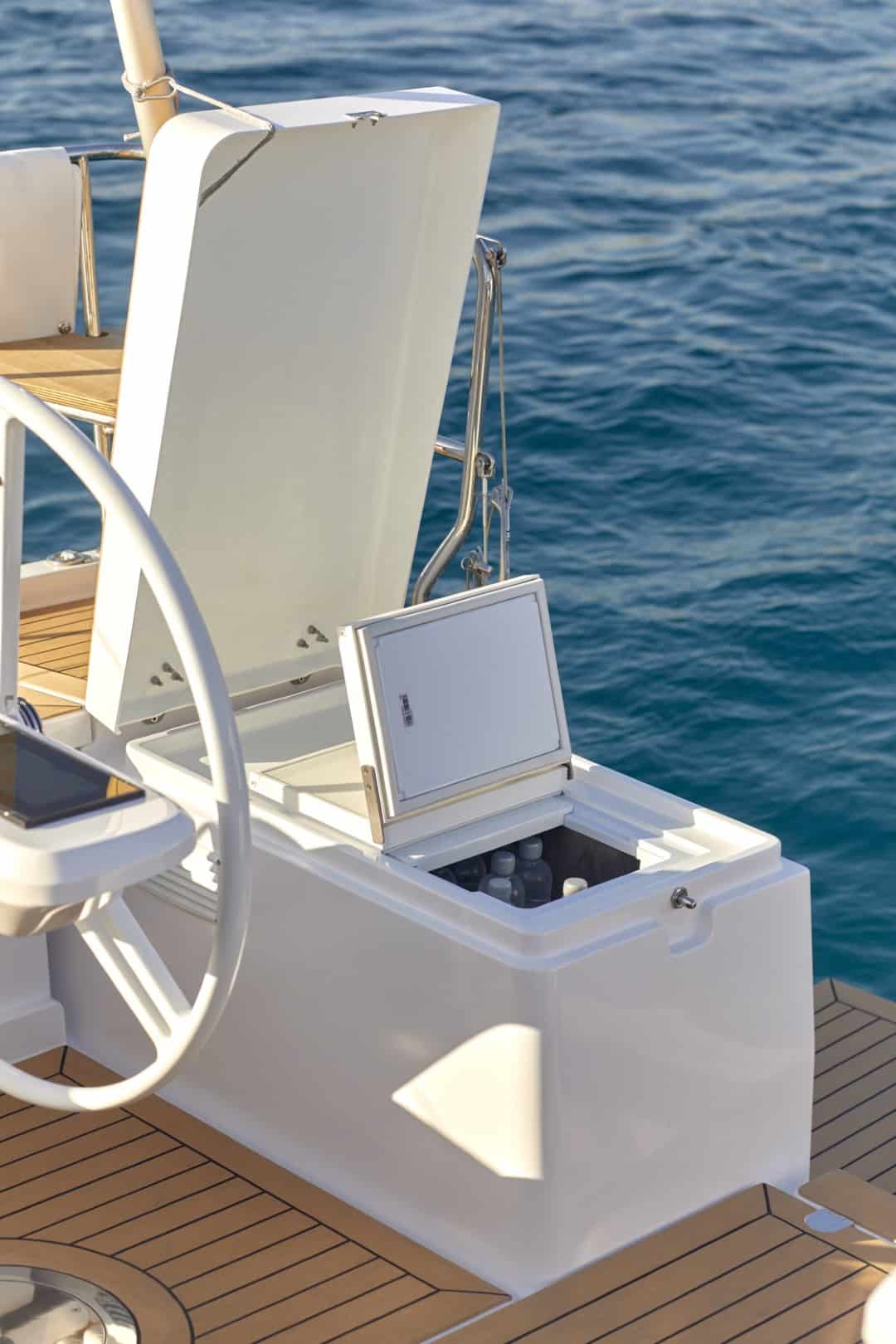 Ein luxuriöses Segelyachtdeck mit eingebauter Kühlbox mit Flaschen neben einem weißen Steuerrad und einem ruhigen blauen Ozean im Hintergrund.