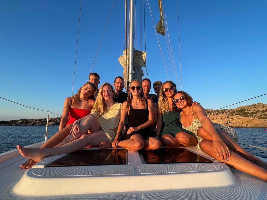 Eine Gruppe von acht Personen, fünf Frauen und drei Männer, sitzt zusammen auf dem Deck eines Katamarans unter einem klaren, sonnigen Himmel. Sie lächeln und posieren für das Foto, mit einer malerischen Kulisse aus ruhigem Wasser und felsigem Gelände in der Ferne während ihres Privattörns.