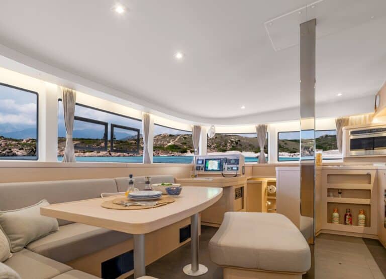 Innenansicht eines modernen Katamarans mit einem gut beleuchteten und geräumigen Wohnbereich mit einem Esstisch, beigen Sofas und mehreren Fenstern, die einen Panoramablick auf die Küstenlandschaft bieten.