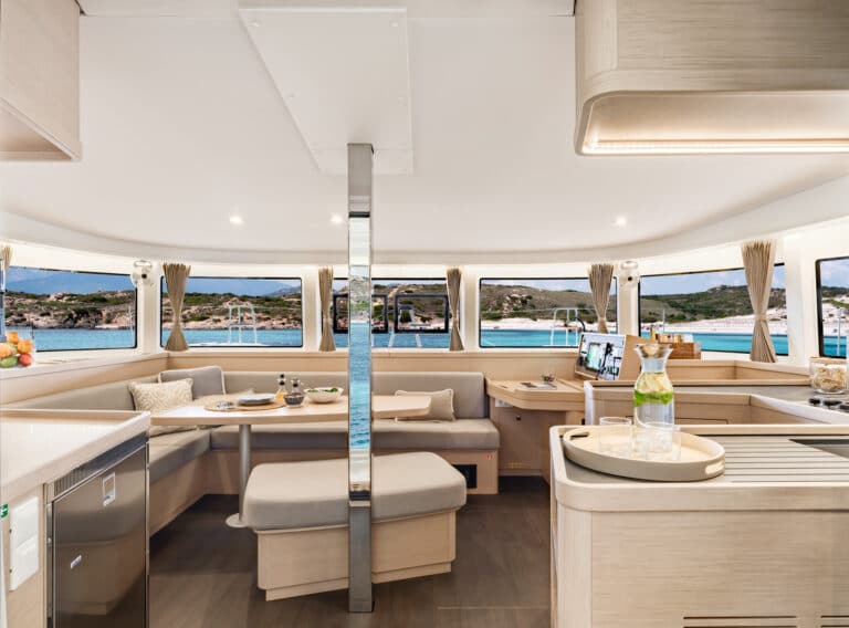 Innenansicht einer modernen Katamaran-Kabine mit einem geräumigen Sitzbereich mit einem zum Essen gedeckten Esstisch, einem Küchenbereich und großen Fenstern mit Blick auf das Meer und die Küste.
