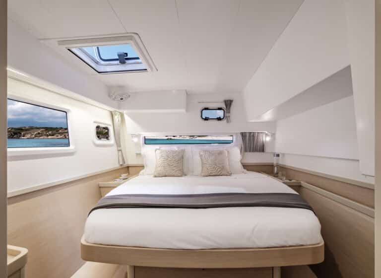 Luxuriöses Yachtkabinen-Interieur mit großem Bett mit weicher Bettwäsche, Deckenbeleuchtung und großen Fenstern mit Meerblick. Modernes und gemütliches Design mit hellen, klaren Tönen, ideal für einen Segelurlaub.