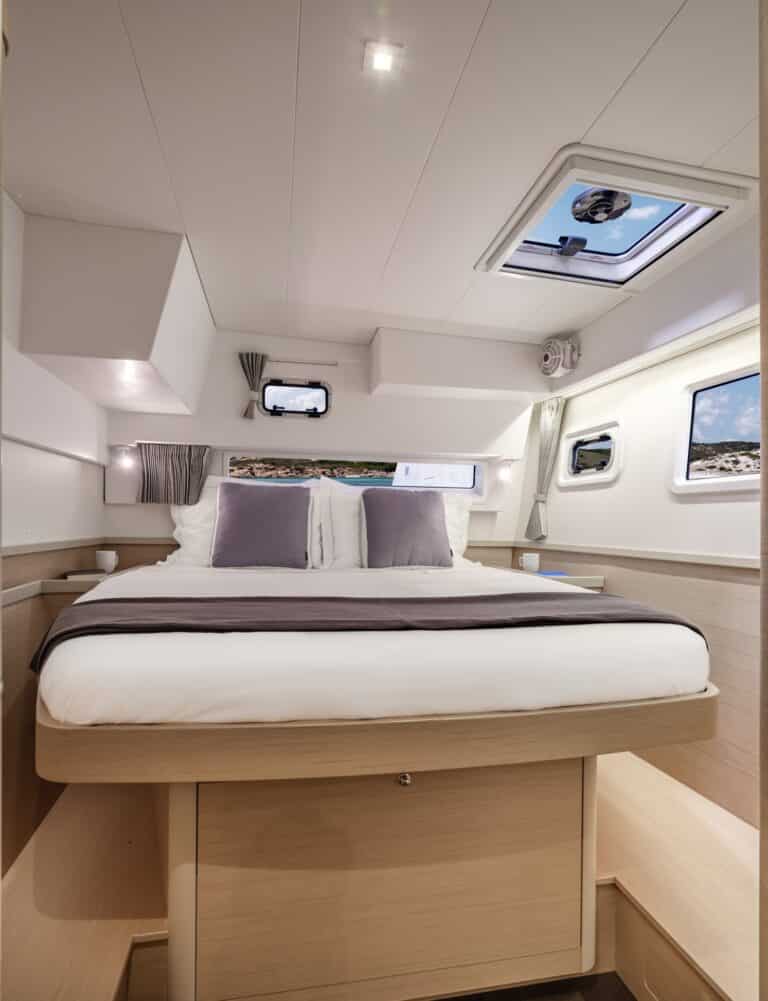 Innenansicht einer modernen Segelyachtkabine mit einem großen Bett mit beiger und grauer Bettwäsche, Holzschränken und mehreren Fenstern für Tageslicht.