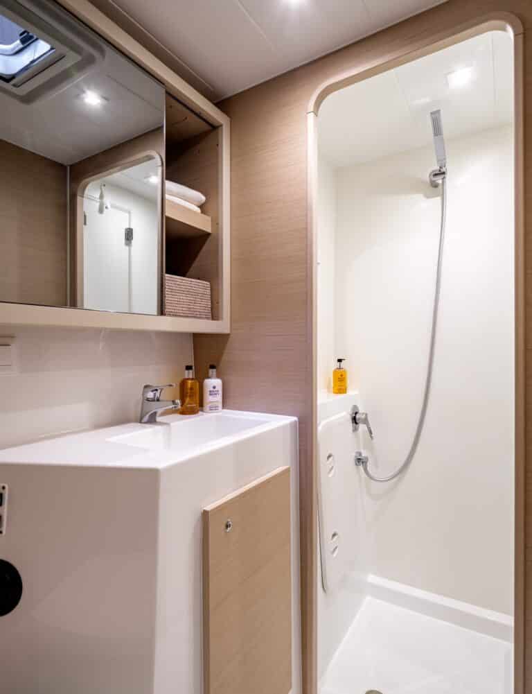 Modernes kleines Badezimmer mit weißem Waschbecken, Spiegelschrank, Glasduschtür und Holzwandpaneelen sowie auf einem Katamaran präsentierten Toilettenartikeln.
