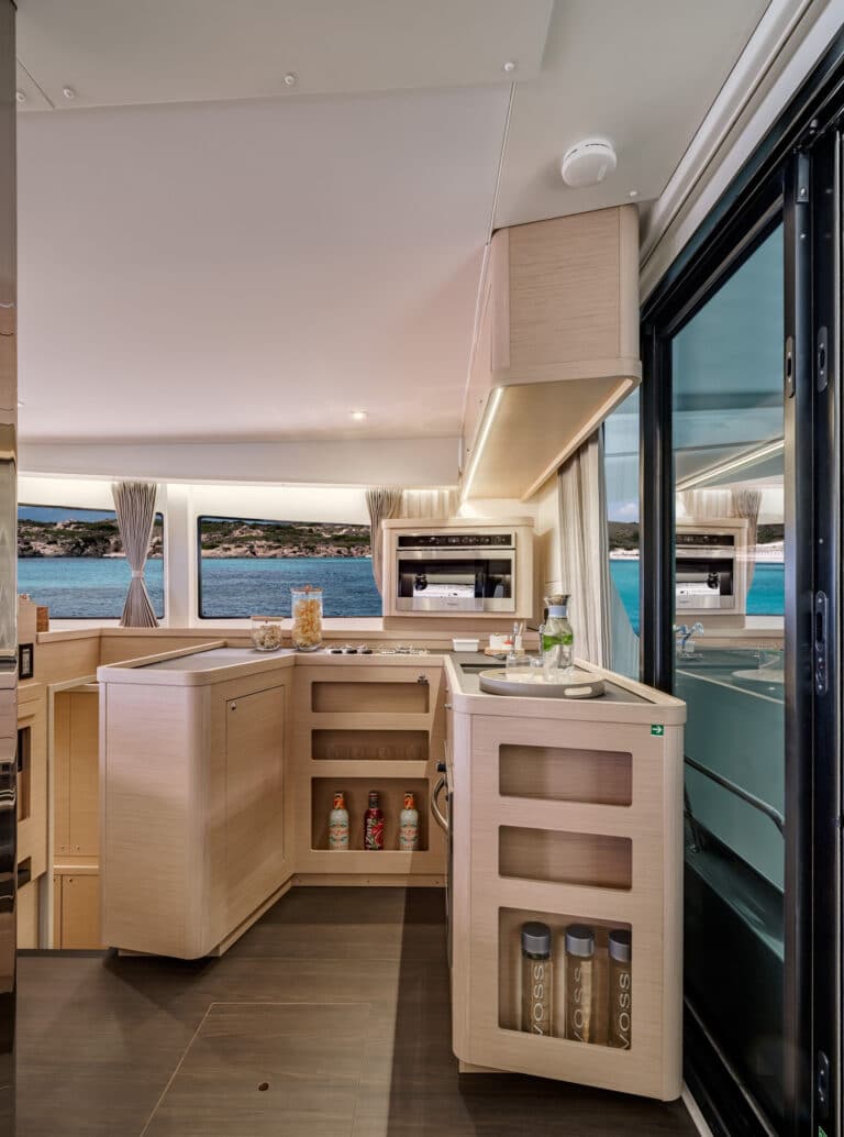 Innenraum einer modernen Yachtküche mit hellen Holzschränken, Einbaugeräten und einem großen Fenster mit Blick auf das Meer während einer Segelreise.