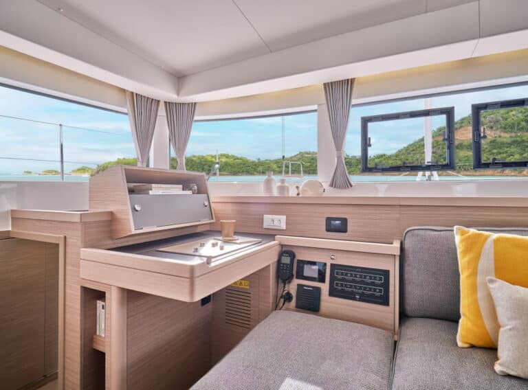 Innenansicht einer modernen Segelyachtkabine mit einer kompakten Küchenzeile mit eleganten Holzeinrichtungen, einer gemütlichen Sitzecke mit einem grauen Sofa und gelben Kissen sowie großen Fenstern mit Blick auf die Küste.