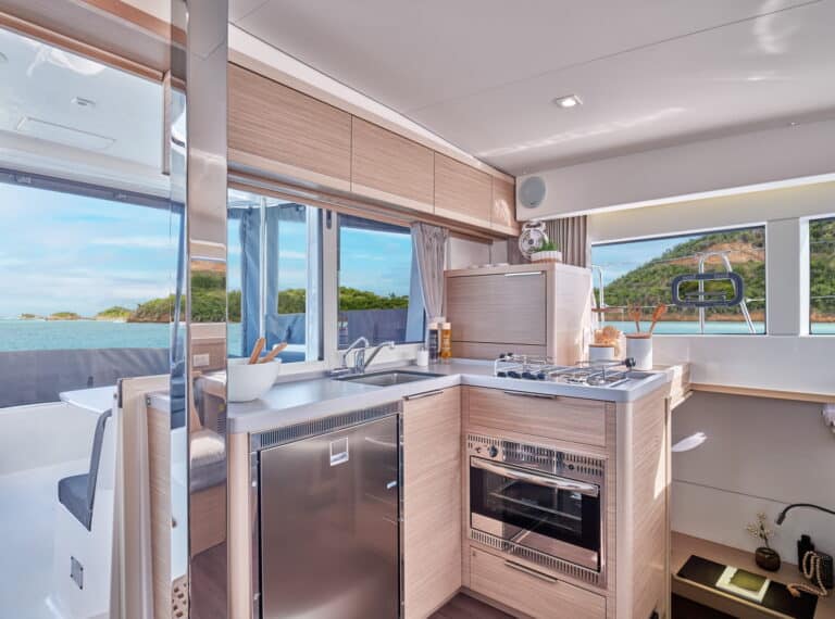 Eine moderne Küche in einer Segelyacht mit Holzschränken, einem Edelstahlkühlschrank, einem Herd und einer Mikrowelle sowie Meerblick durch große Fenster.