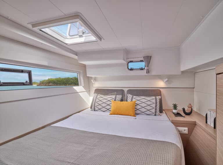 Innenansicht eines modernen Segelyacht-Schlafzimmers mit einem großen Bett mit dekorativen Kissen, Holzschränken und großen Fenstern, die für Tageslicht und eine malerische Aussicht sorgen.