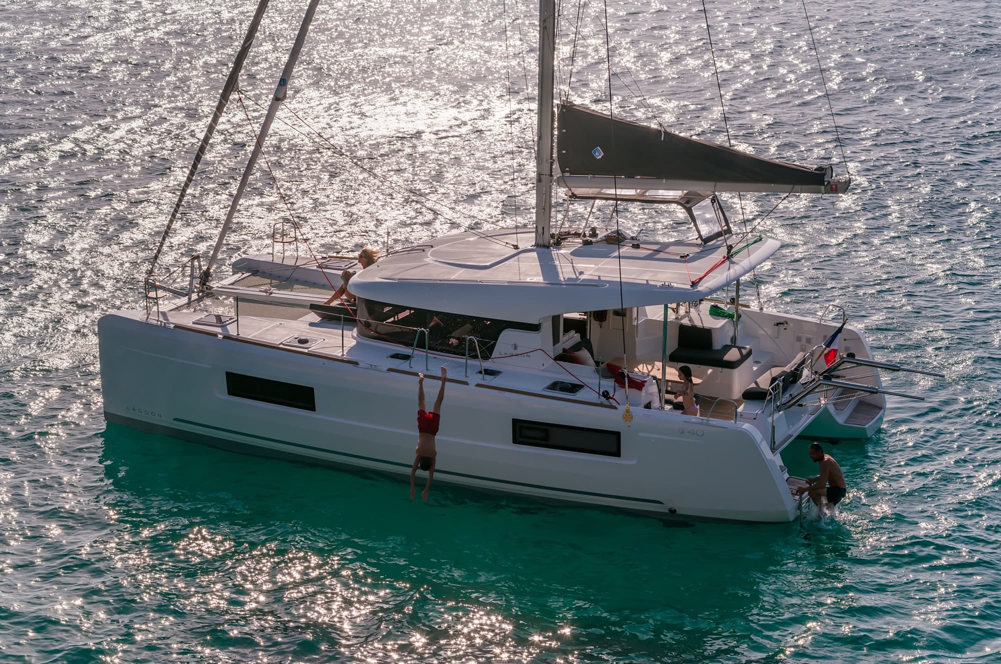 Eine moderne weiße Segelyacht segelt auf glitzerndem türkisfarbenem Wasser, während ein paar Leute sich entspannen und die Sonne genießen. Das Segel ist teilweise entfaltet, was eine sanfte Bewegung suggeriert.