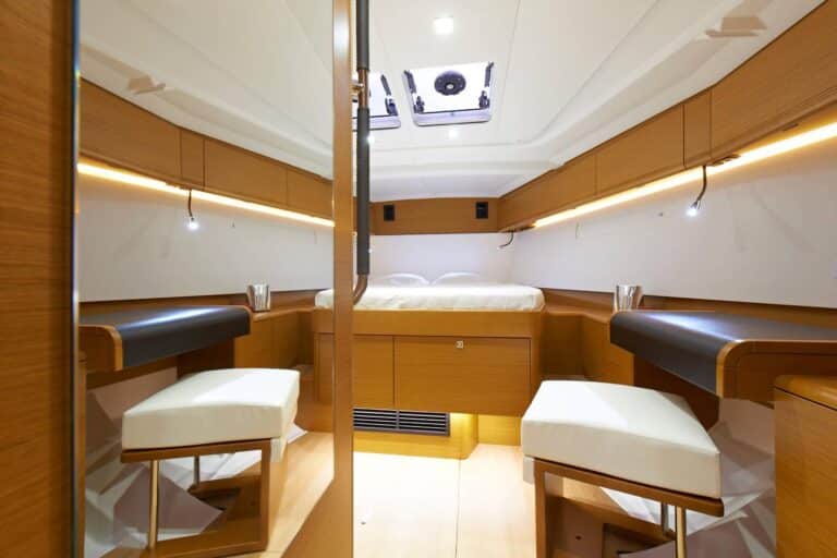 Innenansicht einer modernen Katamarankabine mit einem gemütlichen Bett, zwei kleinen weißen Stühlen, Holzschränken und stimmungsvoller Beleuchtung.