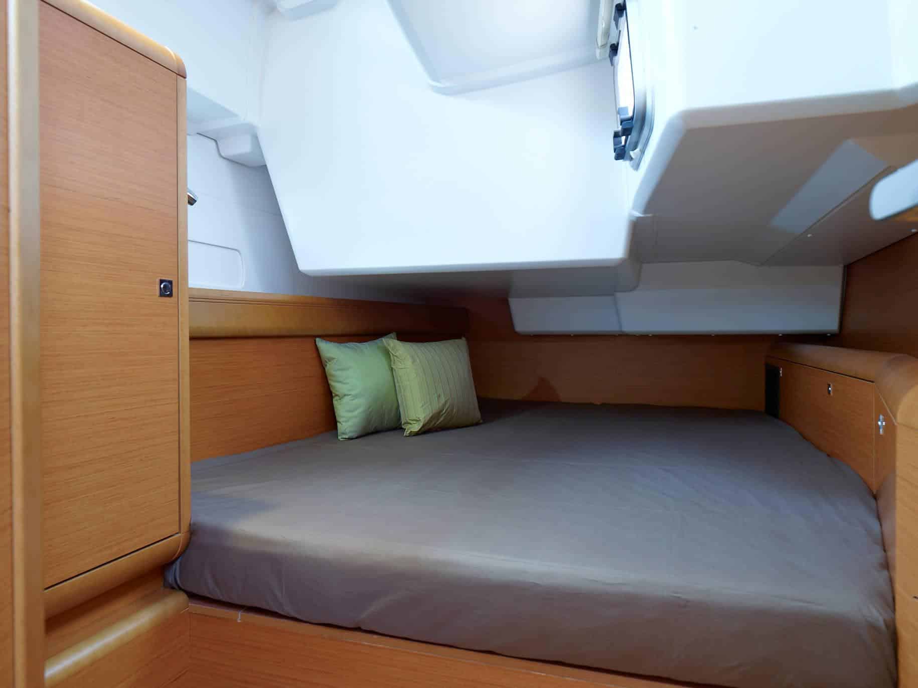 Innenansicht einer Segelyachtkabine mit einem kompakten Bett mit grauer Decke und grünem Kissen, umgeben von Holzpaneelen und einem kleinen Fenster darüber.