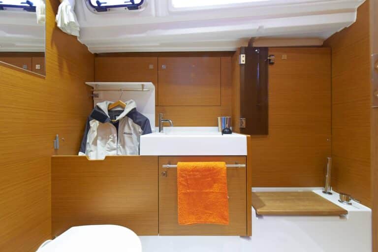 Ein kompaktes Segelyacht-Badezimmer mit Waschbecken, orangefarbenem Handtuch, Spiegel und Holzpaneelen sowie einer ordentlich aufgehängten Kapitänsjacke.