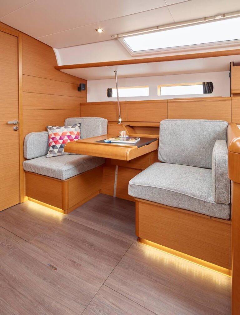Innenansicht einer Katamaran-Kabine mit einem Holztisch, umgeben von grau gepolsterten Sitzbänken und einem einzelnen Sessel, mit warmer Beleuchtung und Holzvertäfelung.