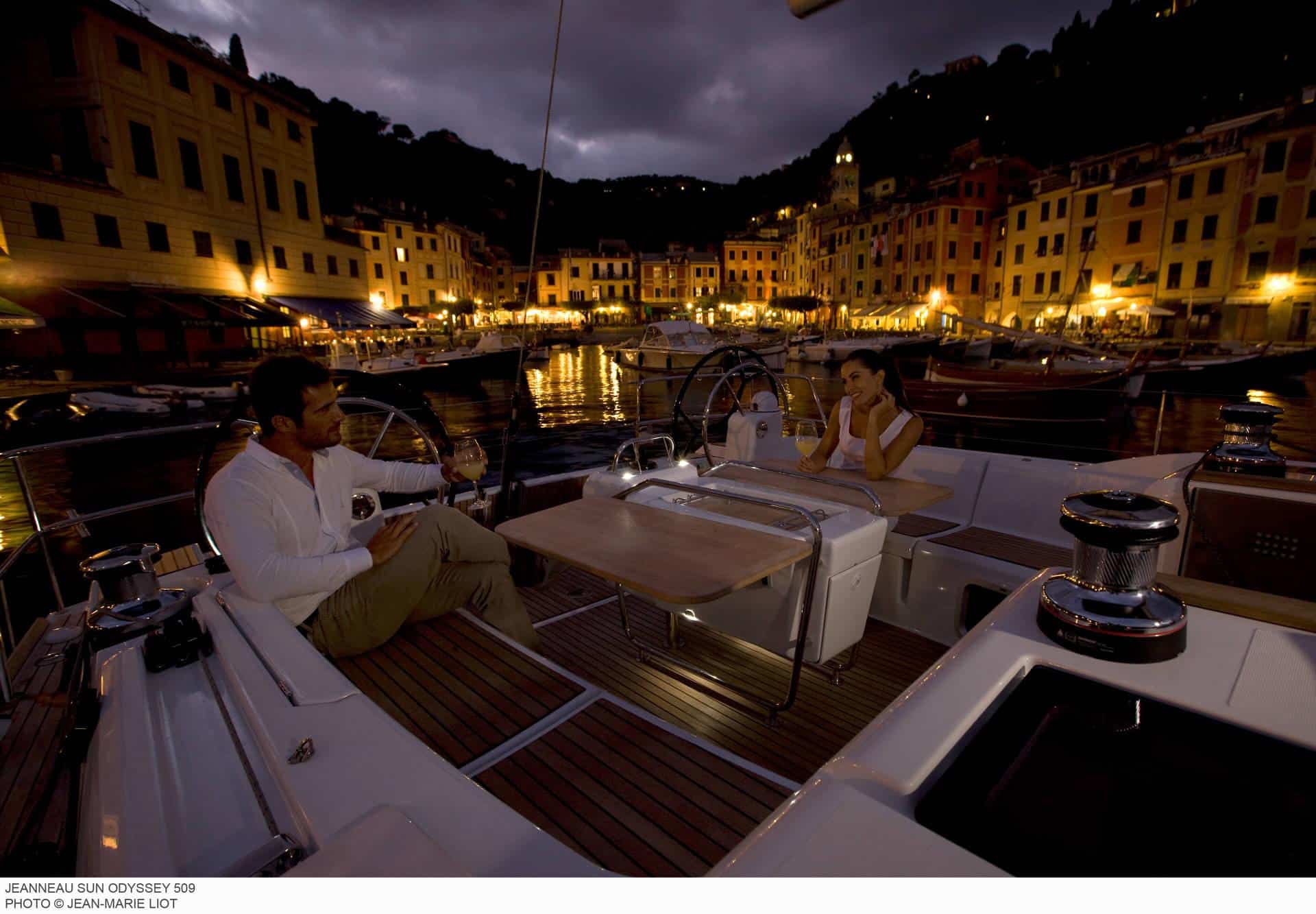 Ein Paar genießt einen romantischen Abend auf einem Katamaran mit einem beleuchteten Küstendorf im Hintergrund. Die Szene wird durch sanftes, stimmungsvolles Licht erhellt, das die ruhige Lage des Yachthafens hervorhebt.