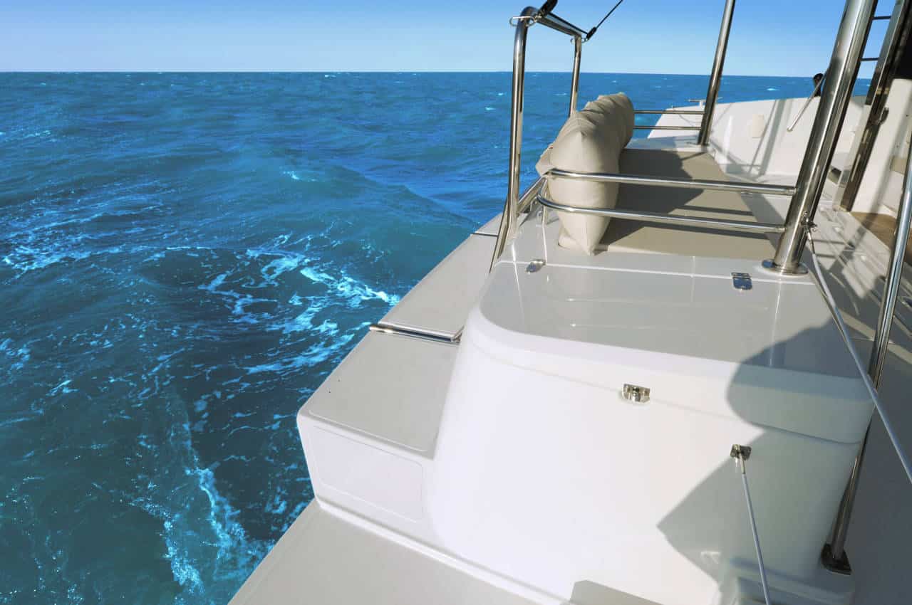 Ein Blick von der Seite eines weißen Bootes auf einem Segeltörn, das auf einem strahlend blauen Ozean unter klarem Himmel segelt, mit Fokus auf das Deck und die Reling des Bootes.