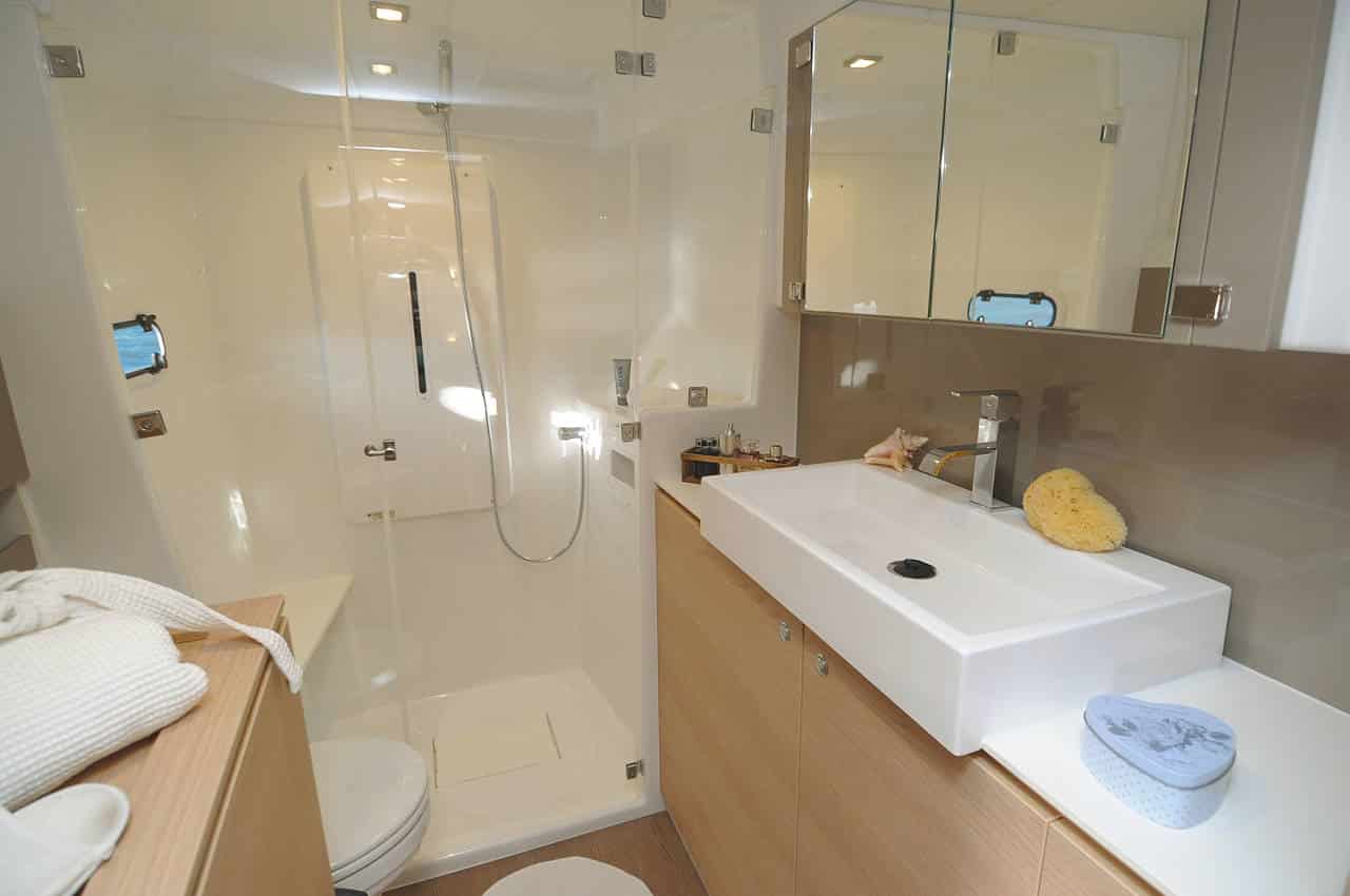 Modernes Badezimmer-Interieur mit gläserner Duschkabine, großem Waschtisch mit Waschbecken, Spiegeln an der Wand und beigefarbenen Fliesen. Handtücher und Toilettenartikel sind ordentlich auf einem Regal mit Segelyacht-Motiv angeordnet.