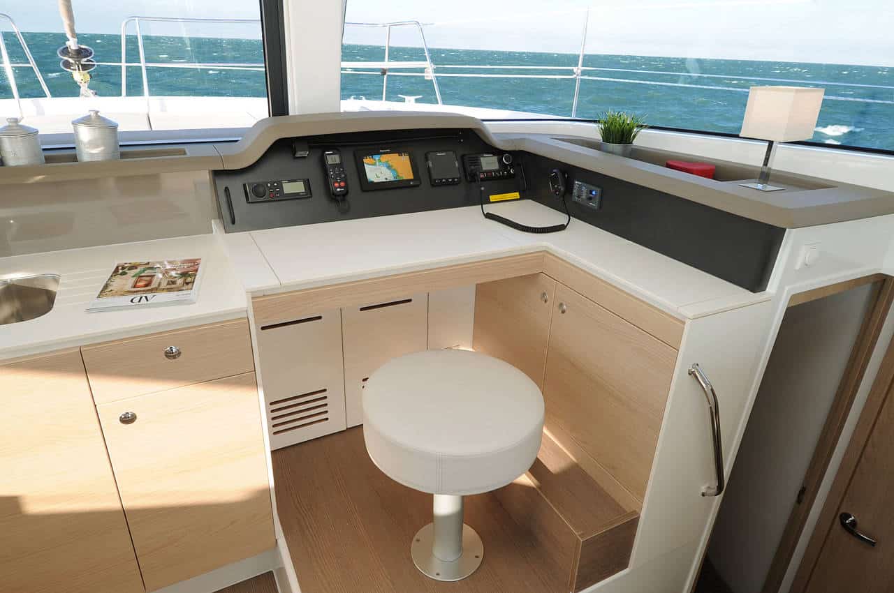 Innenansicht einer modernen Katamarankabine mit Navigationsarmaturenbrett, weißen Schränken, einem kleinen weißen Tisch und einem Hocker. Durch große Fenster hat man einen Blick auf das Meer.