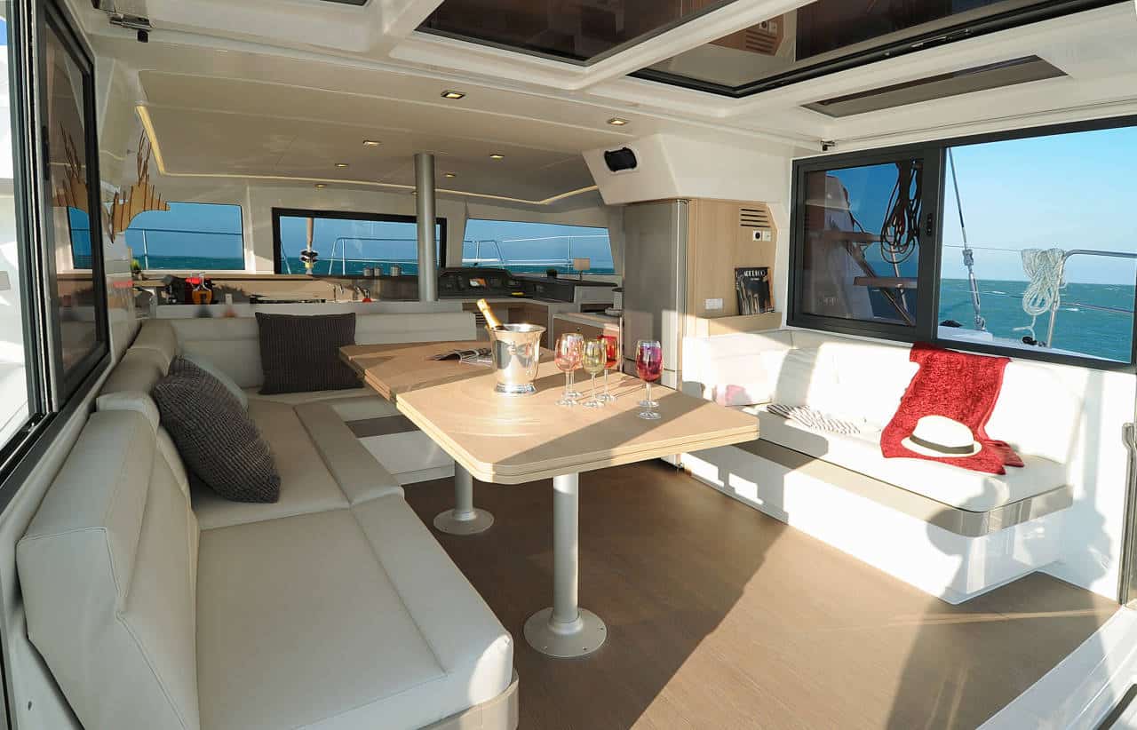 Luxuriöses Segelyacht-Interieur mit geräumigem Loungebereich mit großen Sofas, einem zentralen Tisch und Panoramafenstern mit weitem Meerblick. Erfrischungen werden am Tisch serviert.
