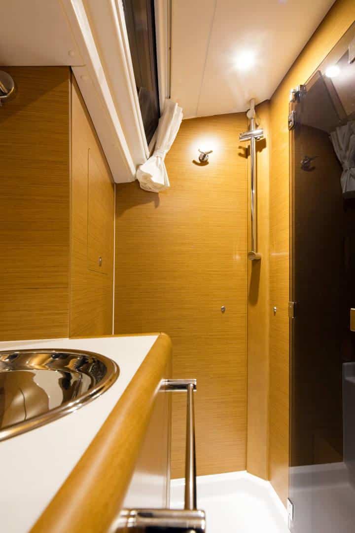 Innenansicht einer kompakten Bootskabine mit einer Holzwand mit Handtuchhalter, einem kleinen runden Waschbecken und einem Teil eines Handlaufs im Vordergrund. Helles Licht erhellt den Raum während der Segelreise.
