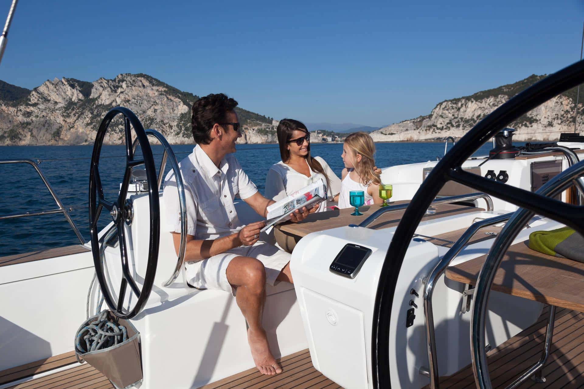 Drei Menschen genießen einen sonnigen Tag auf einem Luxus-Katamaran. Ein Mann steuert das Boot, zwei Frauen sitzen in der Nähe und unterhalten sich. Im Hintergrund ist eine malerische Bergküste zu sehen.