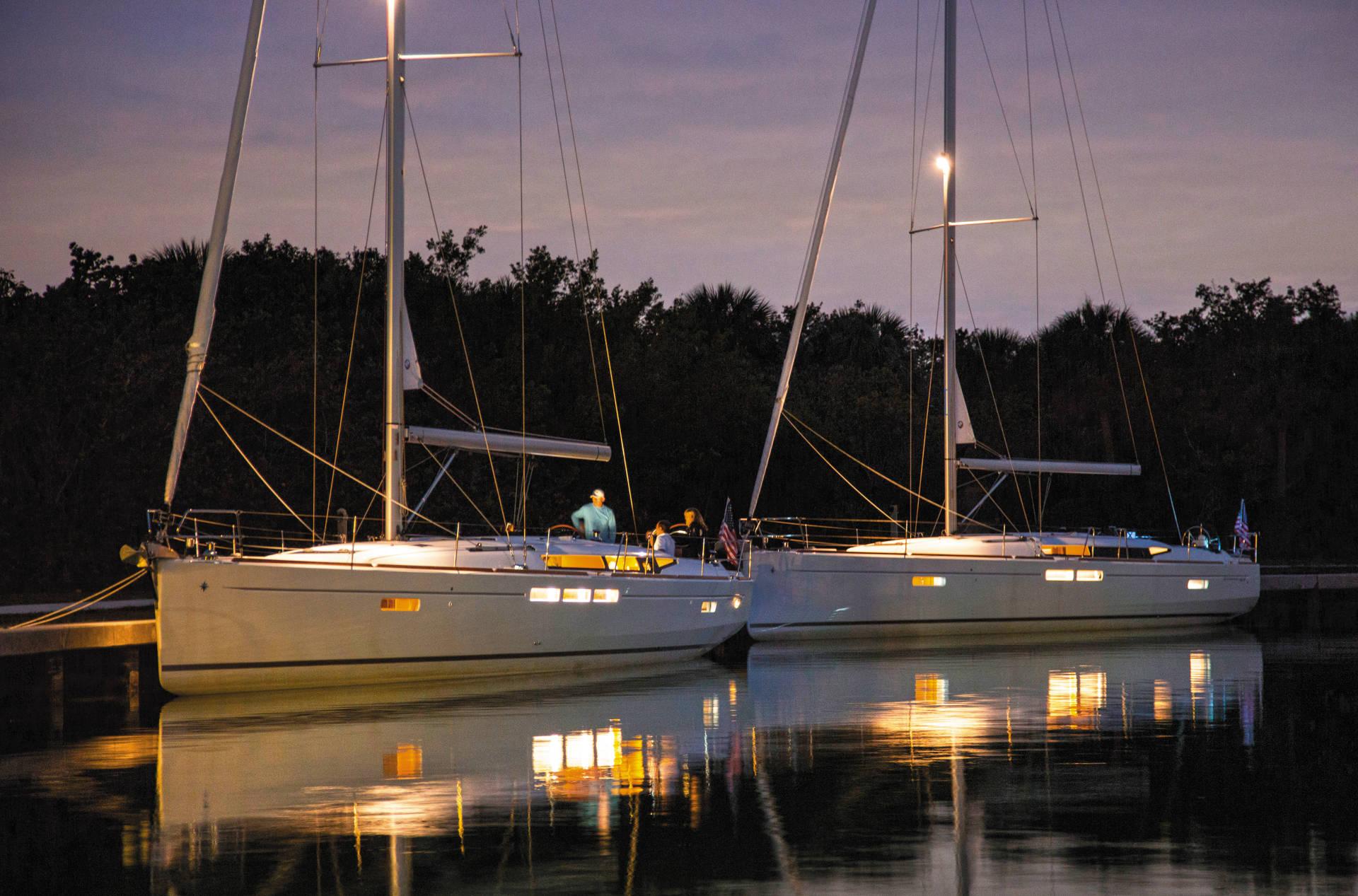 Zwei Segelyachten liegen in der Dämmerung nebeneinander an einem Dock, leuchtende Lichter spiegeln sich im ruhigen Wasser und im Hintergrund ist eine dichte Baumreihe zu sehen.