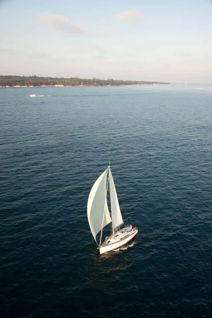 Ein Segelboot gleitet aus der Luftperspektive unter klarem Himmel durch ruhige Gewässer nahe einer Küste. Die Segel des Katamarans sind voll ausgefahren und fangen die Brise ein.