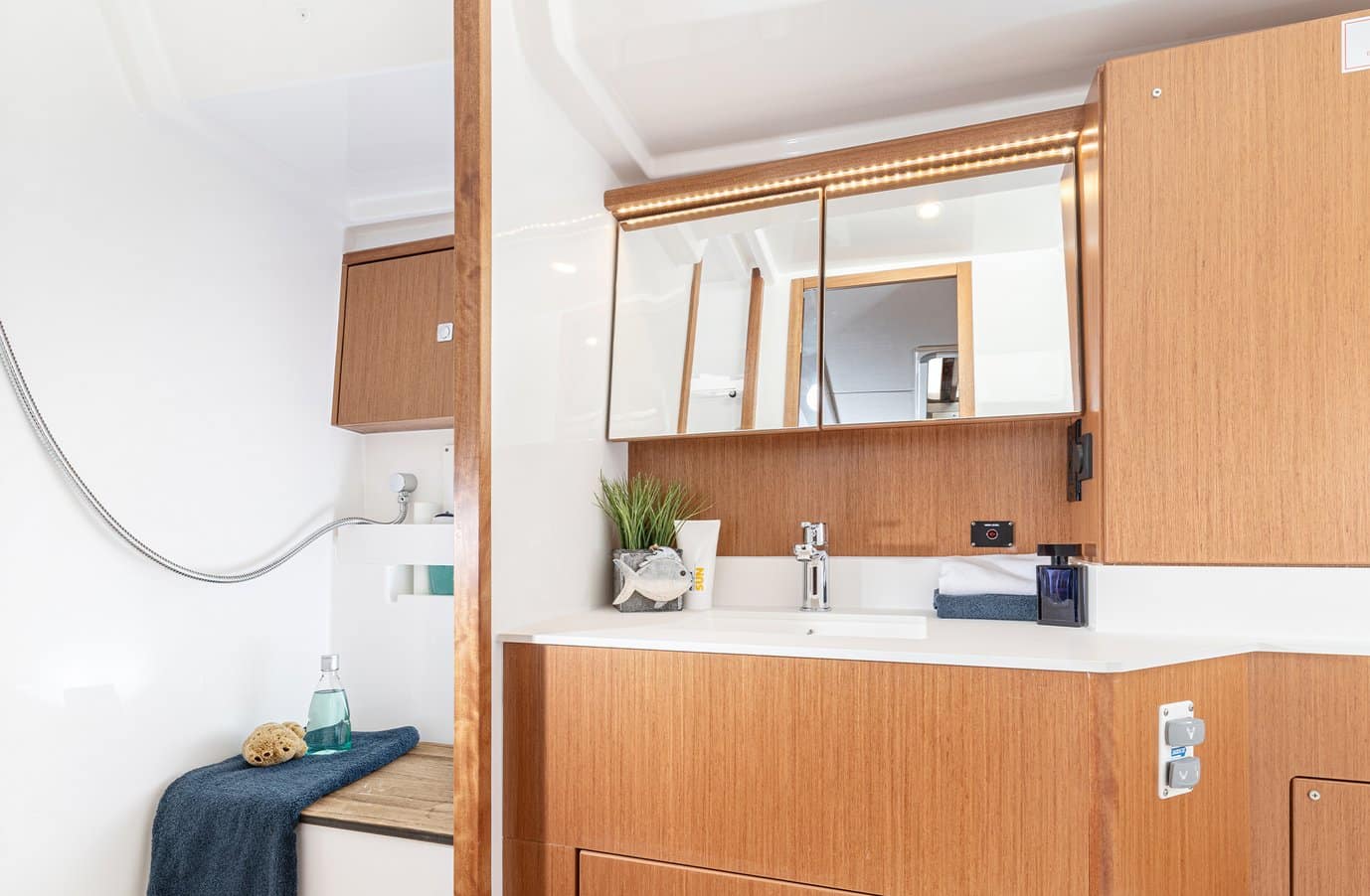 Innenansicht eines kleinen, modernen Badezimmers in einer Segelyacht mit Holzschränken, einem Spiegel mit Schminktischbeleuchtung, einer weißen Arbeitsplatte und minimalistischem Dekor.