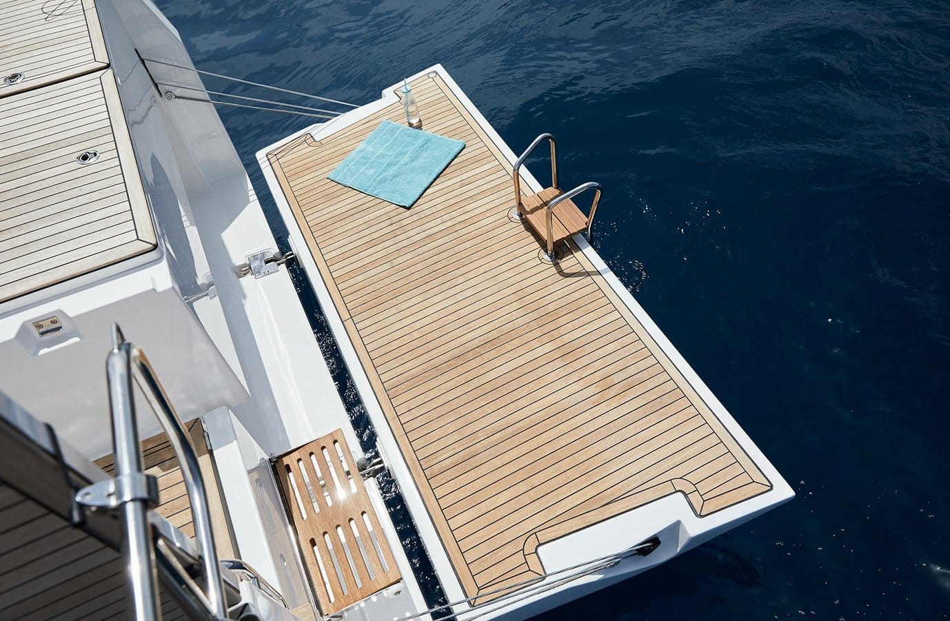 Luftaufnahme des Achterdecks einer Yacht über einem klaren, blauen Meer, mit einem ausgebreiteten Handtuch, einem Holzstuhl und einer ordentlich angeordneten Holzplattform für Segeltörn.