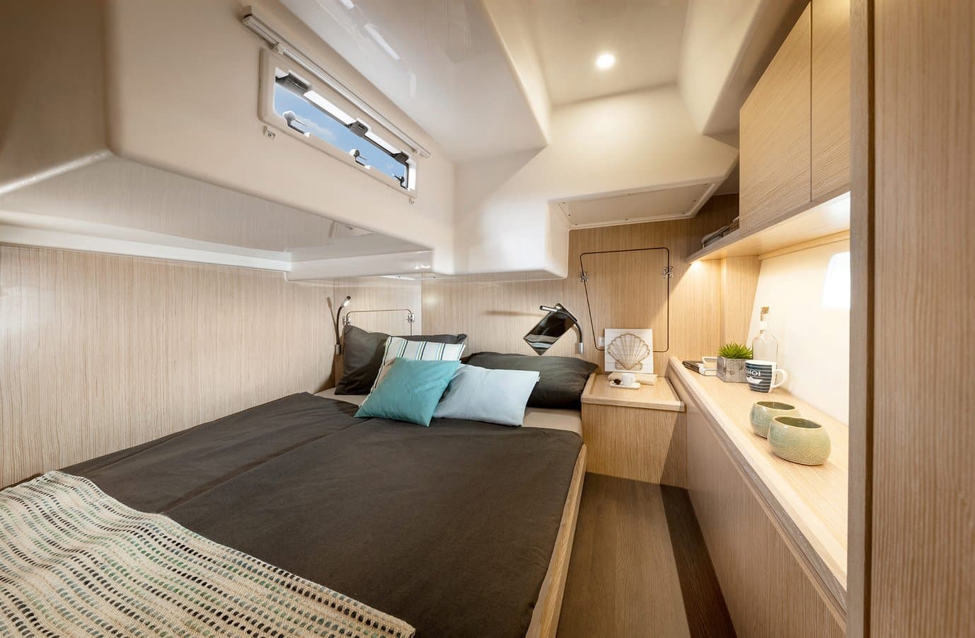 Innenansicht eines modernen Wohnmobils mit gemütlichem Bett mit Kissen, Holzschränken und kleinem Küchenbereich, beleuchtet durch natürliches Licht aus einem Bullauge.