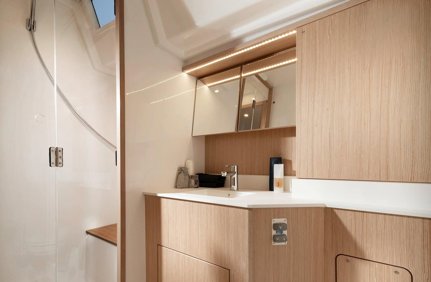 Modernes Badezimmer-Interieur in einer Segelyacht mit hellen Holzpaneelen, einer schlichten weißen Waschtischplatte mit Waschbecken, einem Spiegelschrank und dezenter Beleuchtung.
