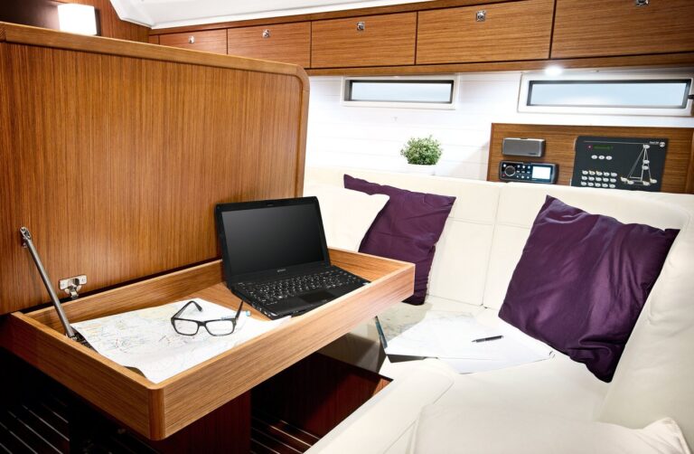 Eine gemütliche Yachtkabine mit einem Holzschreibtisch mit Laptop, Brille und Seekarten. Der umliegende Bereich umfasst plüschige weiße und violette Sitze, holzgetäfelte Wände und moderne Bedienelemente – alles auf einer Segelyacht.