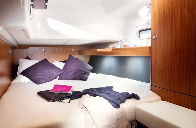 Innenansicht einer Segelyachtkabine mit einem ordentlich gemachten Bett mit violetten Kissen, einem Laptop, einem rosa Notizbuch und einem lässig ausgebreiteten blauen Hemd. Natürliches Licht fällt durch das Bullauge der Kabine ein.