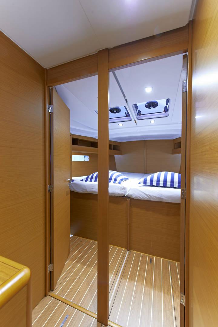 Innenansicht einer Yachtkabine mit zwei Einzelbetten mit blau-weiß gestreifter Bettwäsche, Holzvertäfelung und glattem Boden. Eine Tür im Vordergrund öffnet sich zu dem gemütlichen Abteil, das perfekt für einen Segelurlaub ist.