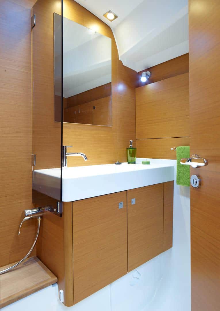 Ein modernes, kompaktes Badezimmer mit Holzschränken, einem weißen Waschbecken und einer weißen Arbeitsplatte sowie einem großen Spiegel. Der Raum ist gut beleuchtet und verfügt über ordentlich aufgehängte grüne Handtücher, perfekt für einen erfrischenden Zwischenstopp während einer Segelreise.