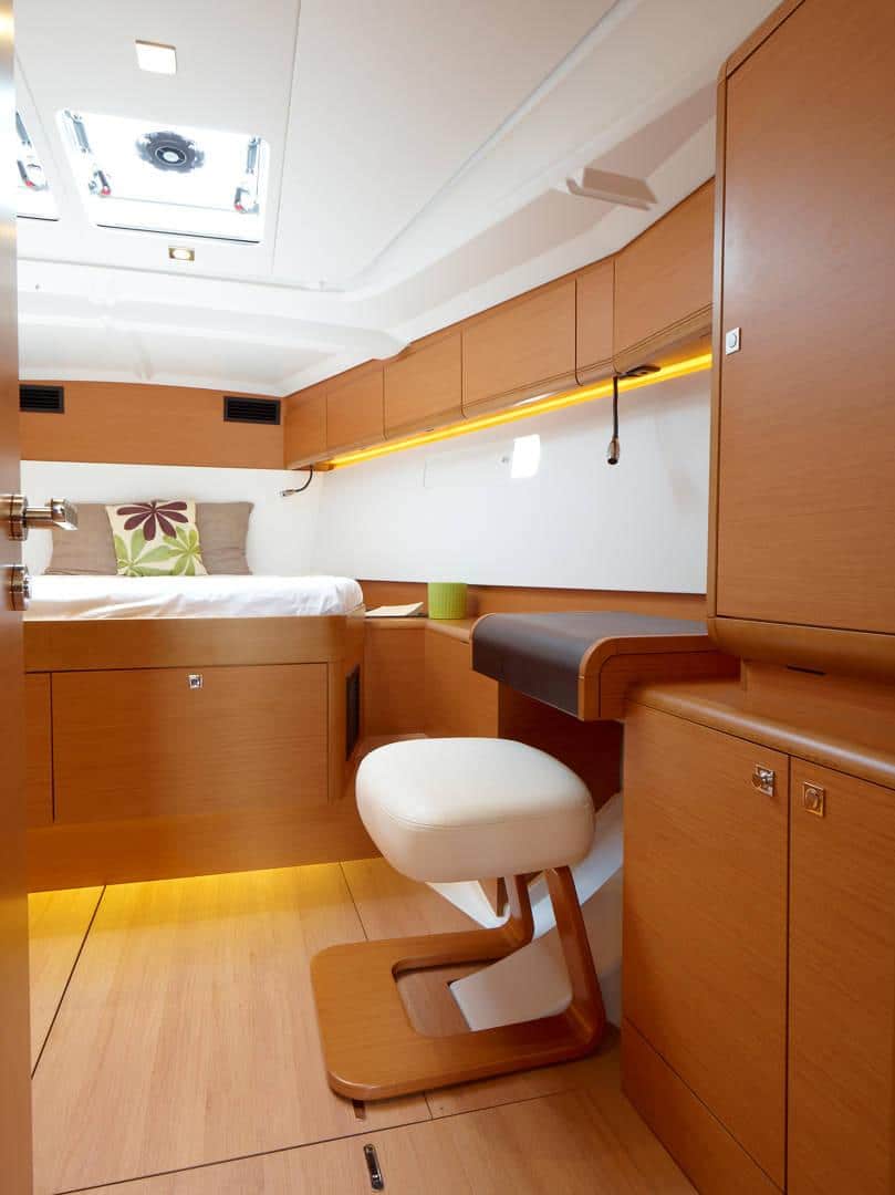 Innenansicht einer luxuriösen Segelyachtkabine mit kompaktem Design, einem Einzelbett, Holzmöbeln, einem kleinen Schreibtisch mit Hocker, warmer Beleuchtung und einem Oberlicht.
