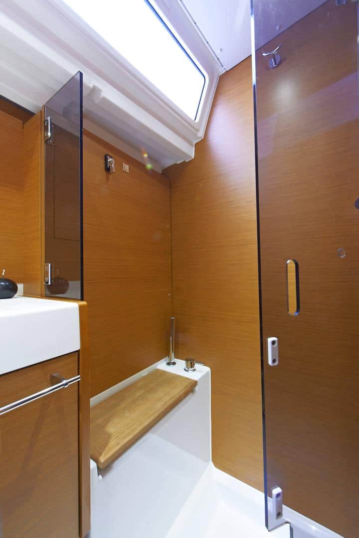 Innenansicht eines modernen Badezimmers mit holzgetäfelten Wänden, einer Duschkabine aus Glas, einem weißen Keramikwaschbecken, einer Holzbank und einem Oberlicht auf einer Segelyacht.