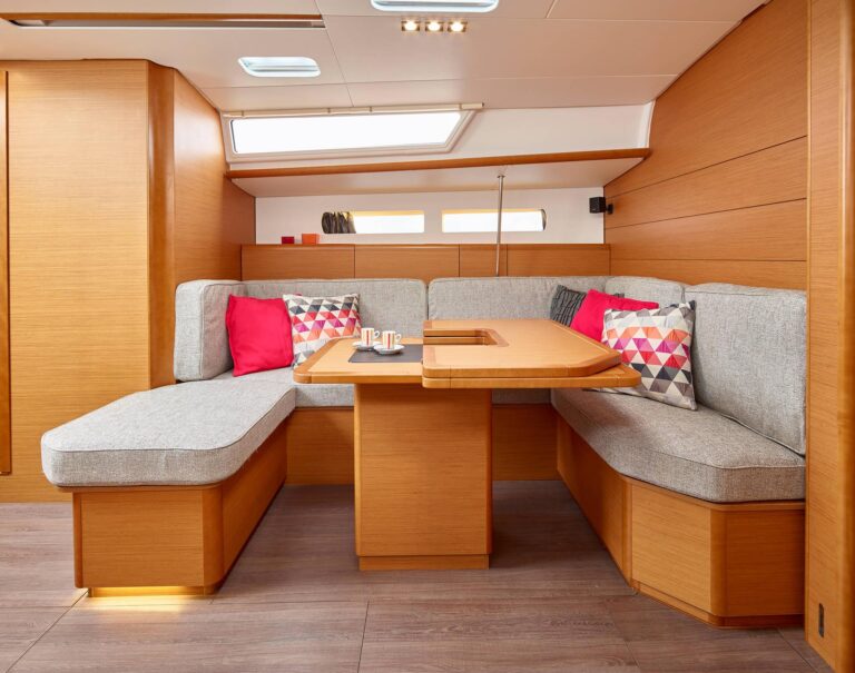 Innenansicht einer für einen Segeltörn entworfenen Bootskabine mit einer U-förmigen Sitzecke mit Holztisch, grauen und gemusterten Kissen sowie Holzpaneelen und -böden.