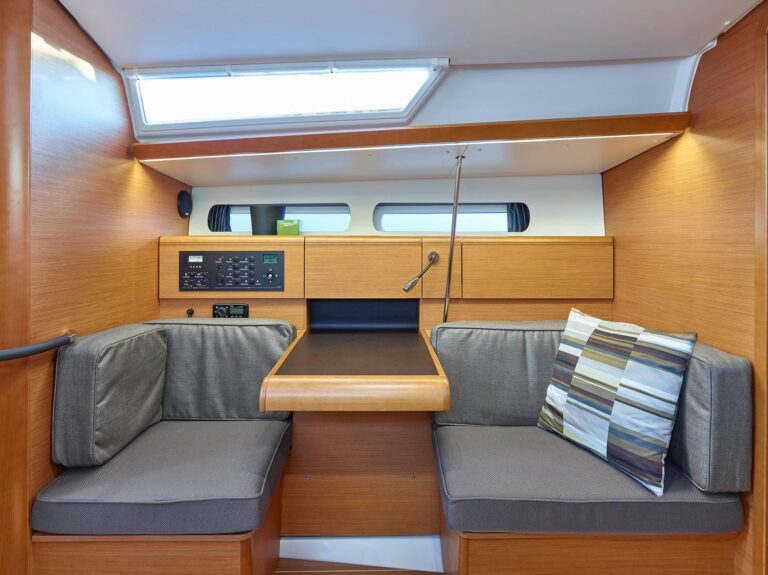 Innenansicht einer modernen Yachtkabine mit zwei grau gepolsterten Sitzbänken, einem Holztisch und Bedienfeldern, mit warmer Holzvertäfelung und natürlichem Licht von oben. Ideal für einen Segelurlaub auf einem Katamaran.